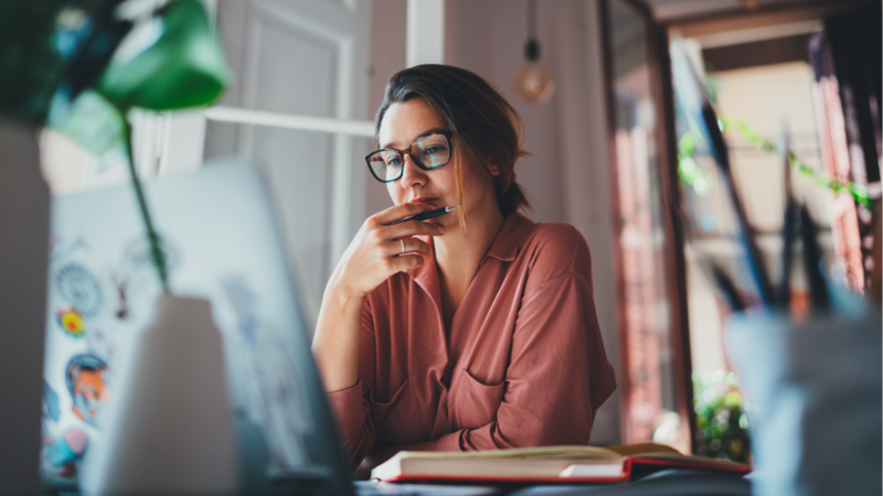 A woman wearing glasses sits at a desk, focused on her laptop while working or studying.