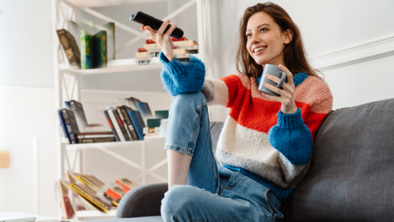 A woman sitting on a couch, casually holding a remote control in her hand, looking relaxed and engaged.