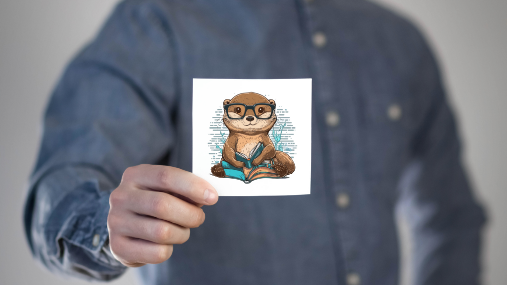 Man holding a kiss-cut sticker of a cartoon otter with glasses holding a book, printed on a square, white backing paper.