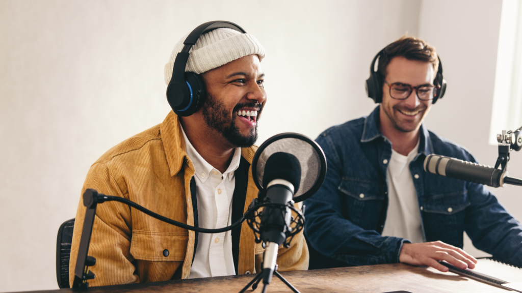 Two men wearing headphones are seated at a desk, engaged in conversation near a microphone.