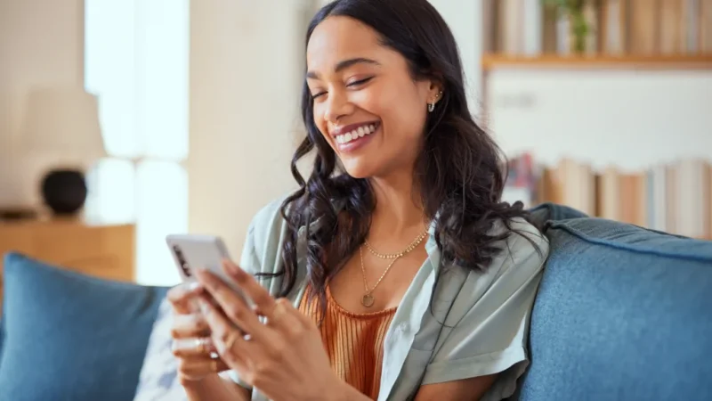 A smiling woman with dark hair is looking at a smartphone in her hand.