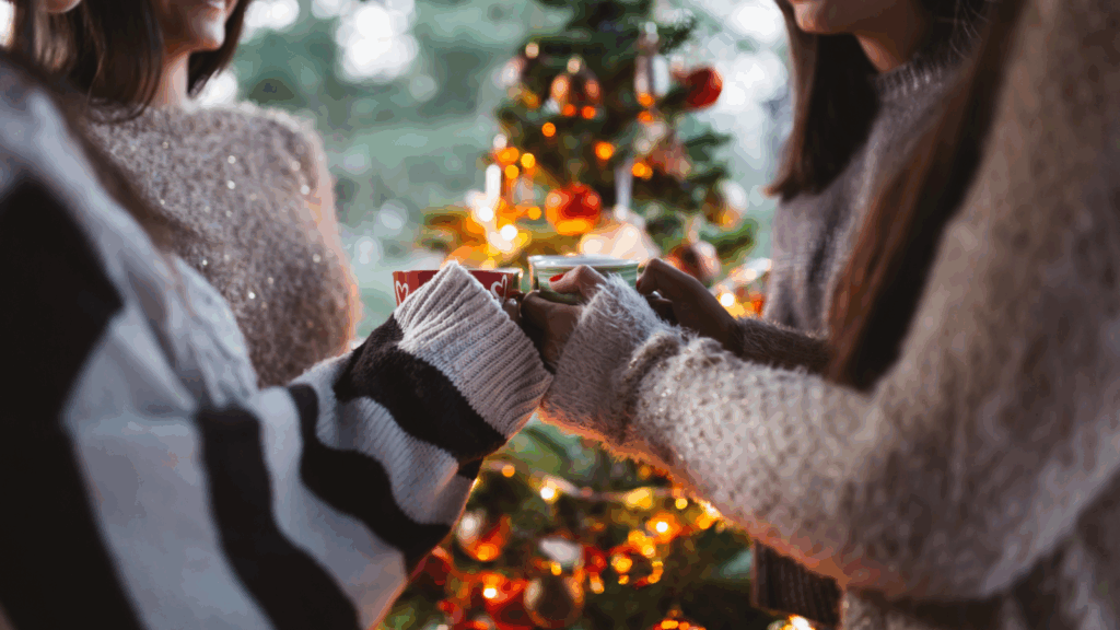Three people in cozy sweaters are exchanging mugs near a decorated Christmas tree.