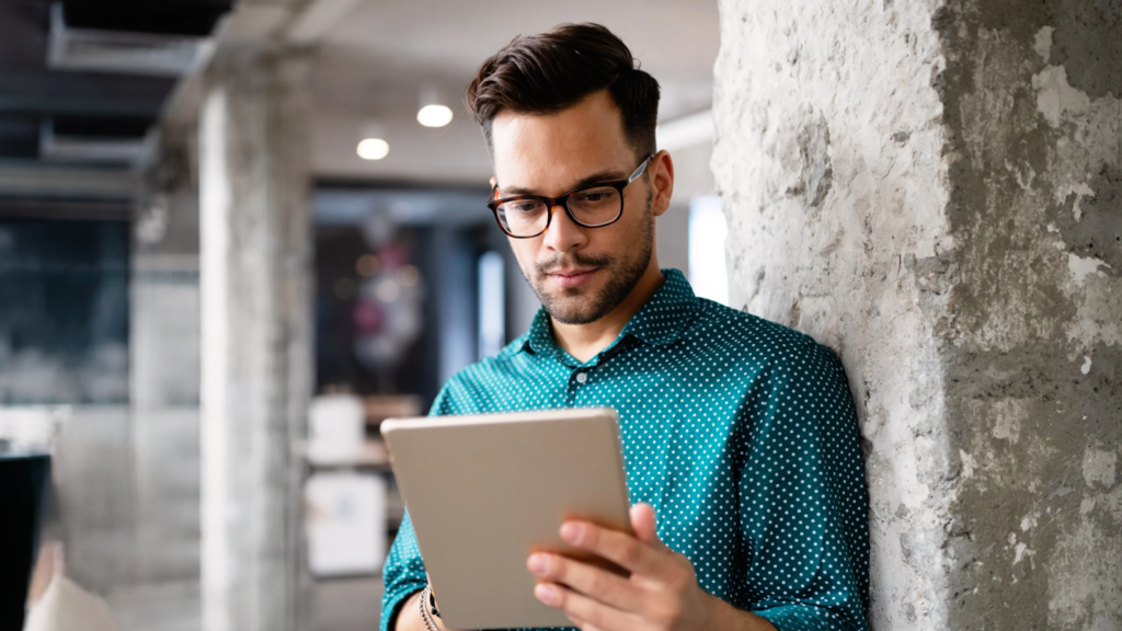 A man in glasses and a teal polka-dot shirt leans against a concrete pillar, reading a comparison of Shopify vs Etsy on a tablet.