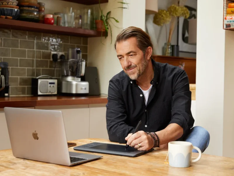 A man in a black shirt works on a digital tablet and a laptop, searching for kidswear’ business ideas.