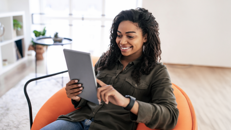 A smiling woman sitting in an orange chair is using a tablet.