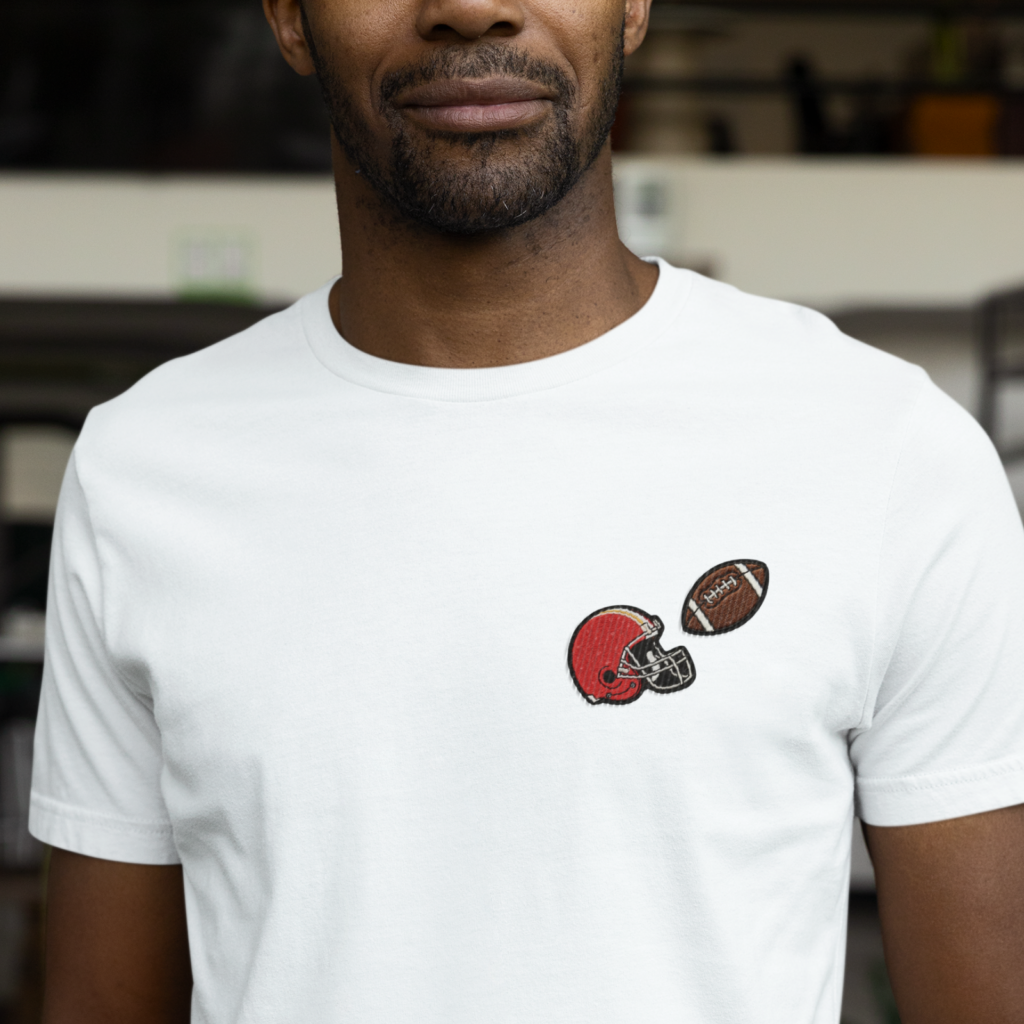 A man wearing a white t-shirt with a small embroidered red football helmet and a brown football.