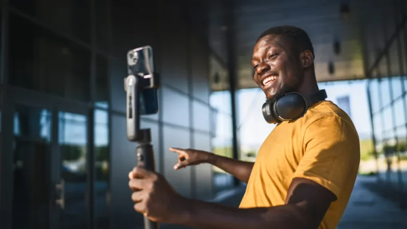 A smiling man with headphones in a yellow shirt holds a smartphone on a selfie stick, gesturing and looking at the camera.