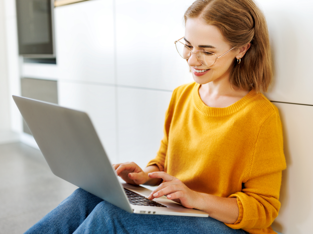 A woman sitting on the floor, working on her laptop, brainstorming ideas for her Shopify t-shirt business.