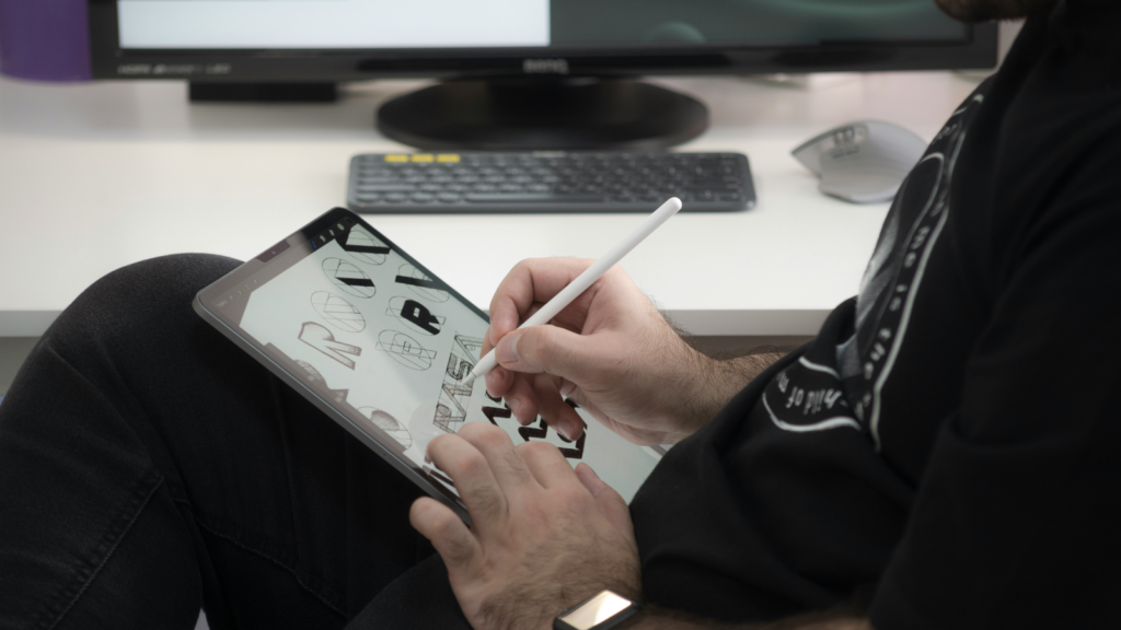 A man in a black hoodie sitting in an office char and creating product designs using a tablet.
