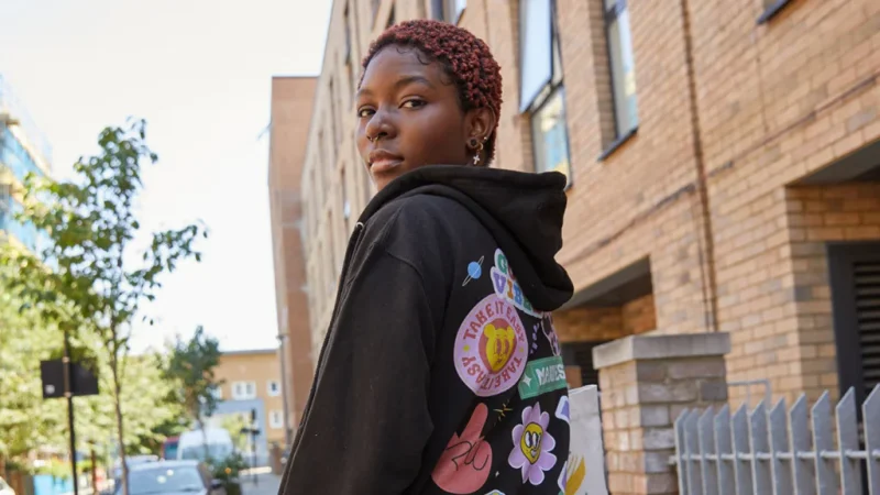 A backshot of a woman wearing a black hoodie with various illustrations printed on the back, looking sideways at the camera.