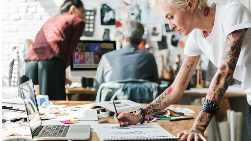 An artist sketching in a notebook while looking at her laptop screen for reference.