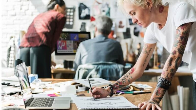 An artist sketching in a notebook while looking at her laptop screen for reference.