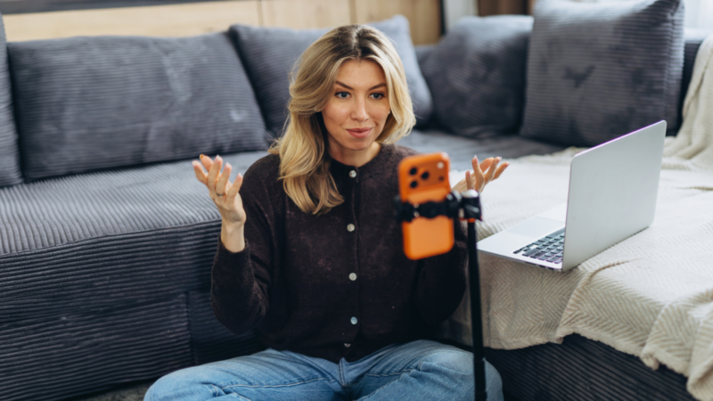 A woman is filming herself using an orange smartphone on a tripod, with a laptop nearby.
