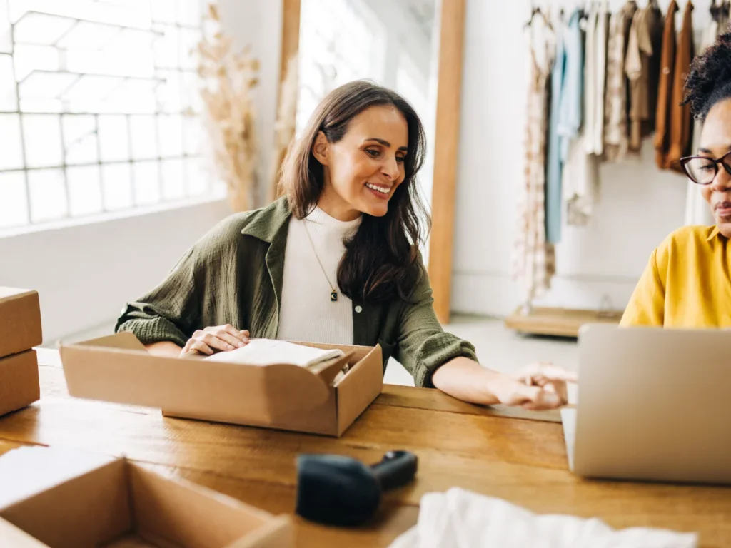 A woman preparing a delivery box for shipment while conversing with another woman.