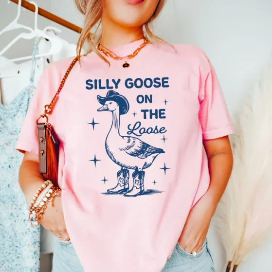 A woman in a pink t-shirt with a cartoon of a goose wearing a cowboy hat and boots. The text reads "Silly Goose on the Loose".