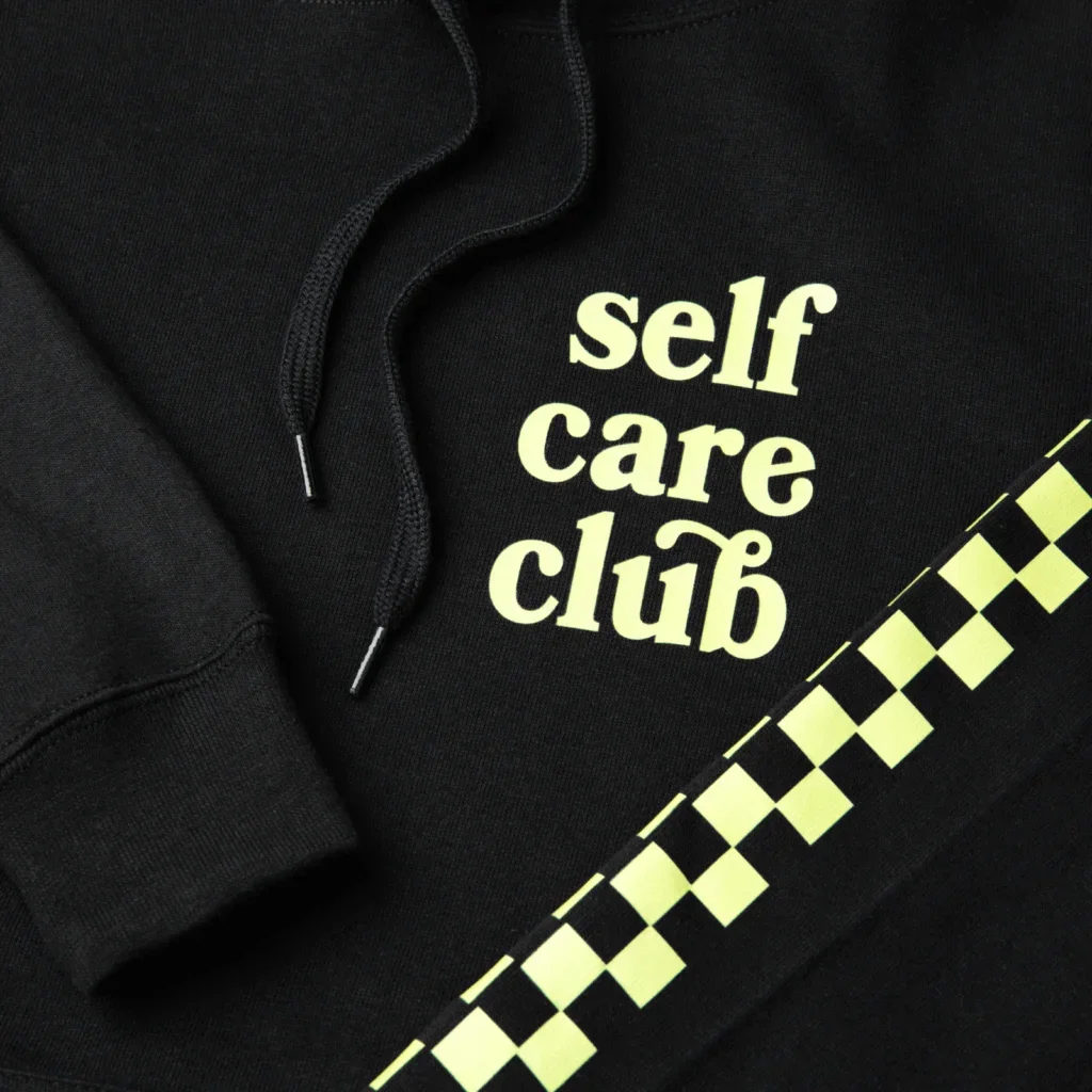 Black hoodie with "self care club" printed in light yellow. Features black drawstrings and a neon green checkered stripe.