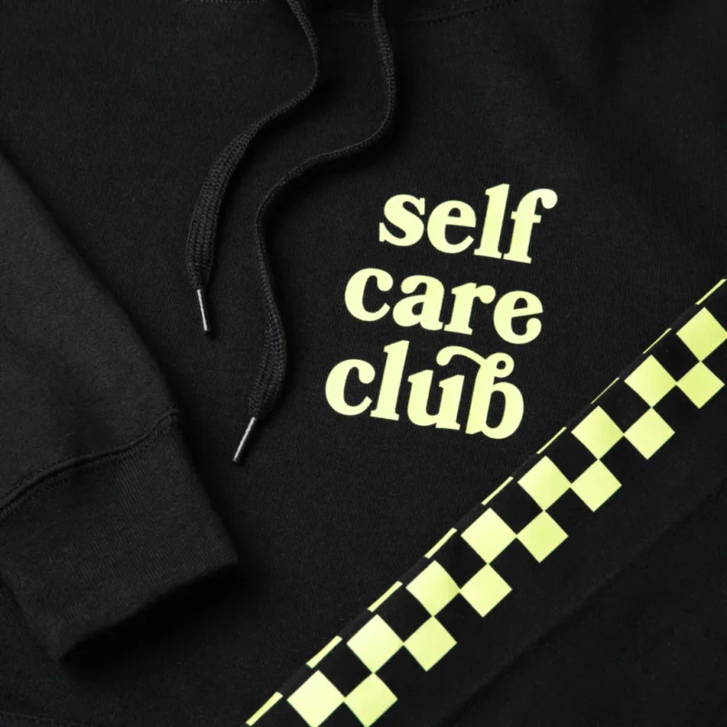 Black hoodie with "self care club" printed in light yellow. Features black drawstrings and a neon green checkered stripe.