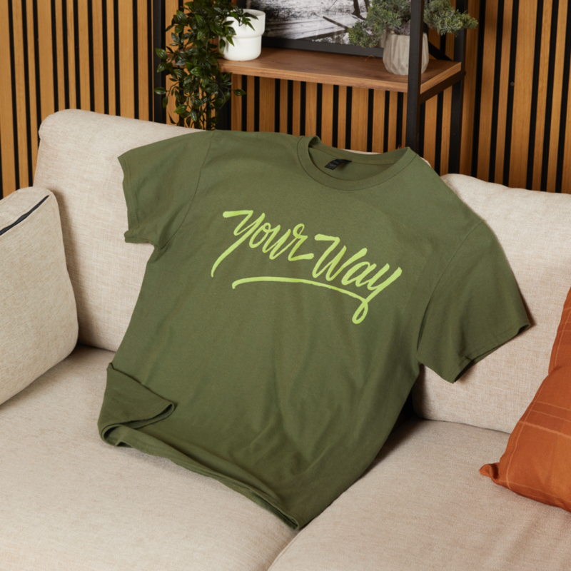 Green t-shirt with "Your Way" in yellow cursive font rests on a beige sofa. A small plant and decor items sit on a wooden shelf behind it.