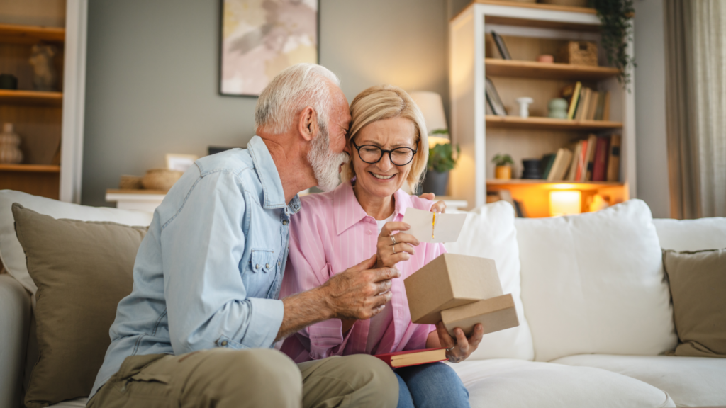 An older couple sits comfortably on a couch, beaming at a gift box and reading a card.