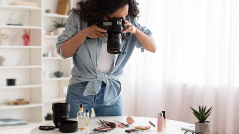 A woman is taking pictures of makeup products with a DSLR camera. White shelves with various items in the background.