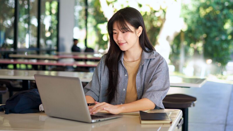 A smiling young woman with black hair, wearing a striped shirt, is looking at a laptop, learning how to be successful on Etsy.