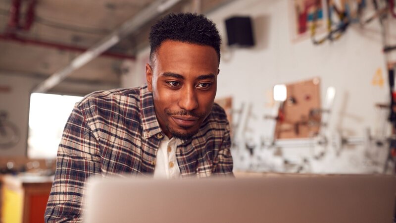 A man in a flannel shirt is focused on a laptop screen, reading how to promote a t-shirt business.