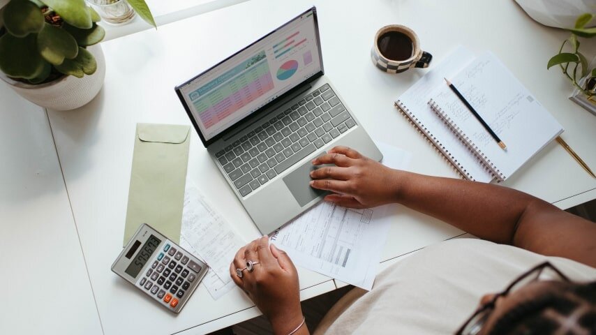 A human sitting next to laptop, doing some calculations, with cup of coffee, some notepads and calculator.
