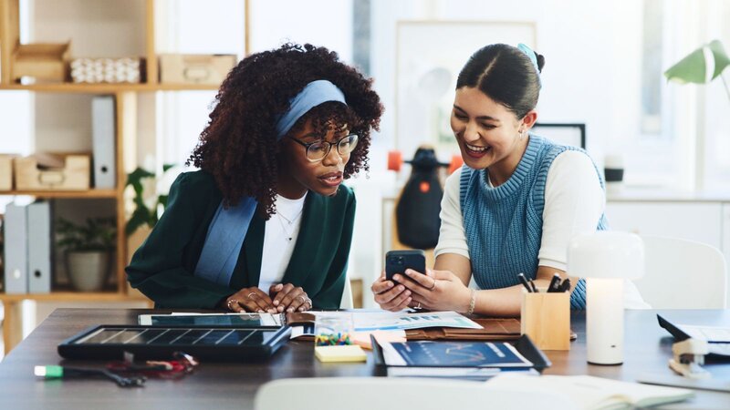 Two women in an office setting share a laugh while looking at a smartphone. The desk is scattered with papers and stationery.