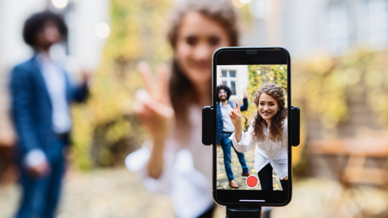 A woman smiling, flashing a peace sign in focus on a smartphone screen, with a blurred background of a man and autumn leaves, creating a cheerful ambiance.
