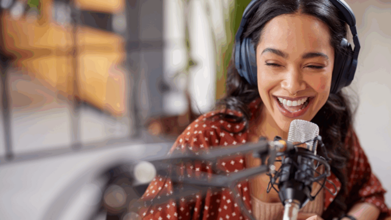 A smiling woman wearing headphones speaks into a microphone.