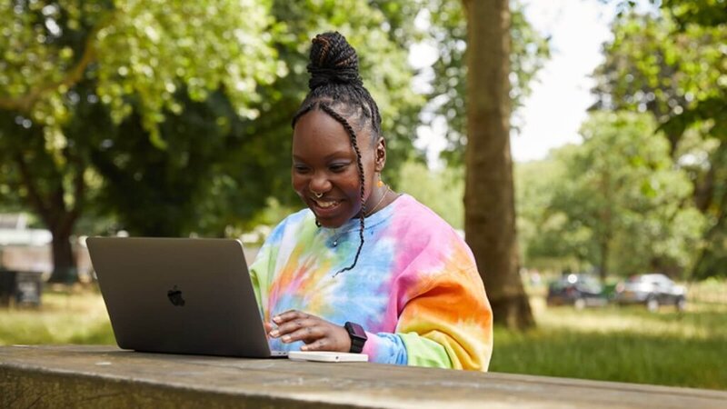 A woman sitting at a bench in a park, working on her laptop.