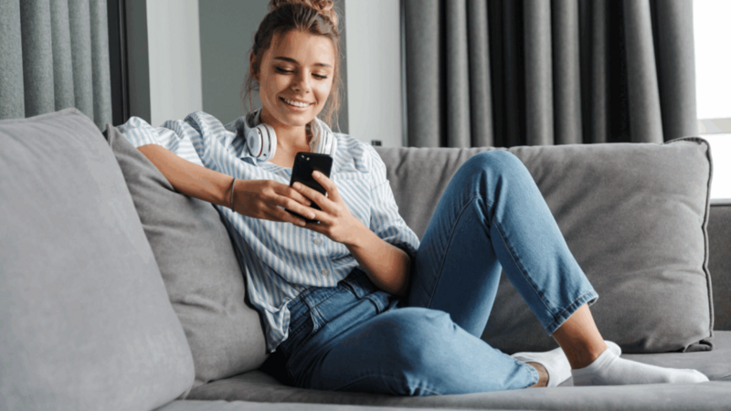 A smiling young woman is sitting on a couch and using a mobile phone to learn how to use TikTok hashtags.