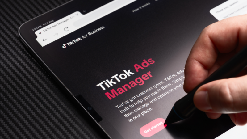 A tablet displays the "TikTok Ads Manager" page. A hand holding a stylus is poised to tap a "Get Started" button. 