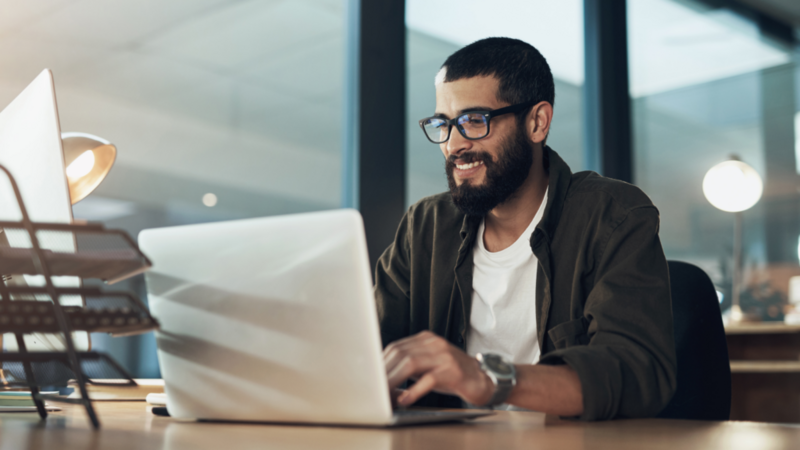 A smiling man in glasses sitting in front of a laptop, behind a desk.