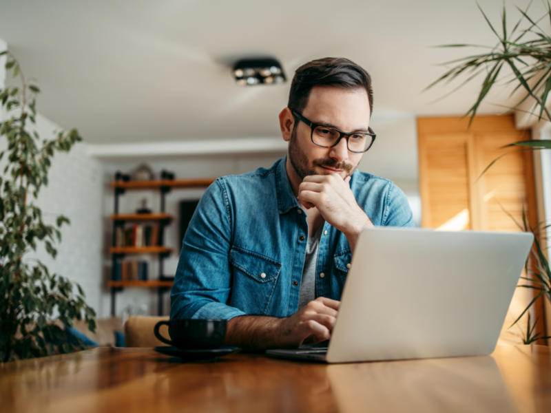 A man in glasses sits at a table, focused on his laptop, ready to sell his stickers online.