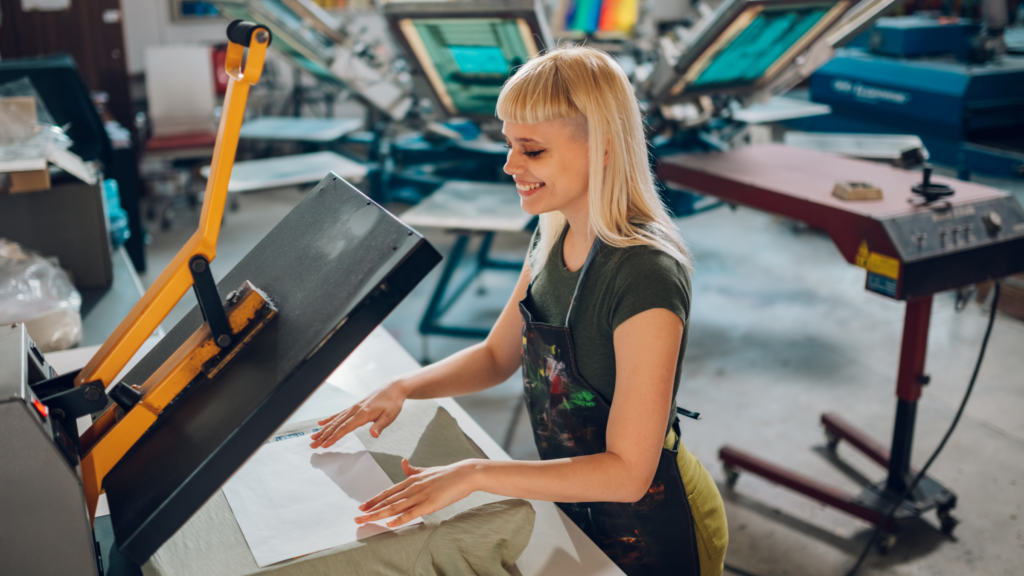 A woman diligently works on a DTF printer, surrounded by tools and materials in a bright, organized studio.