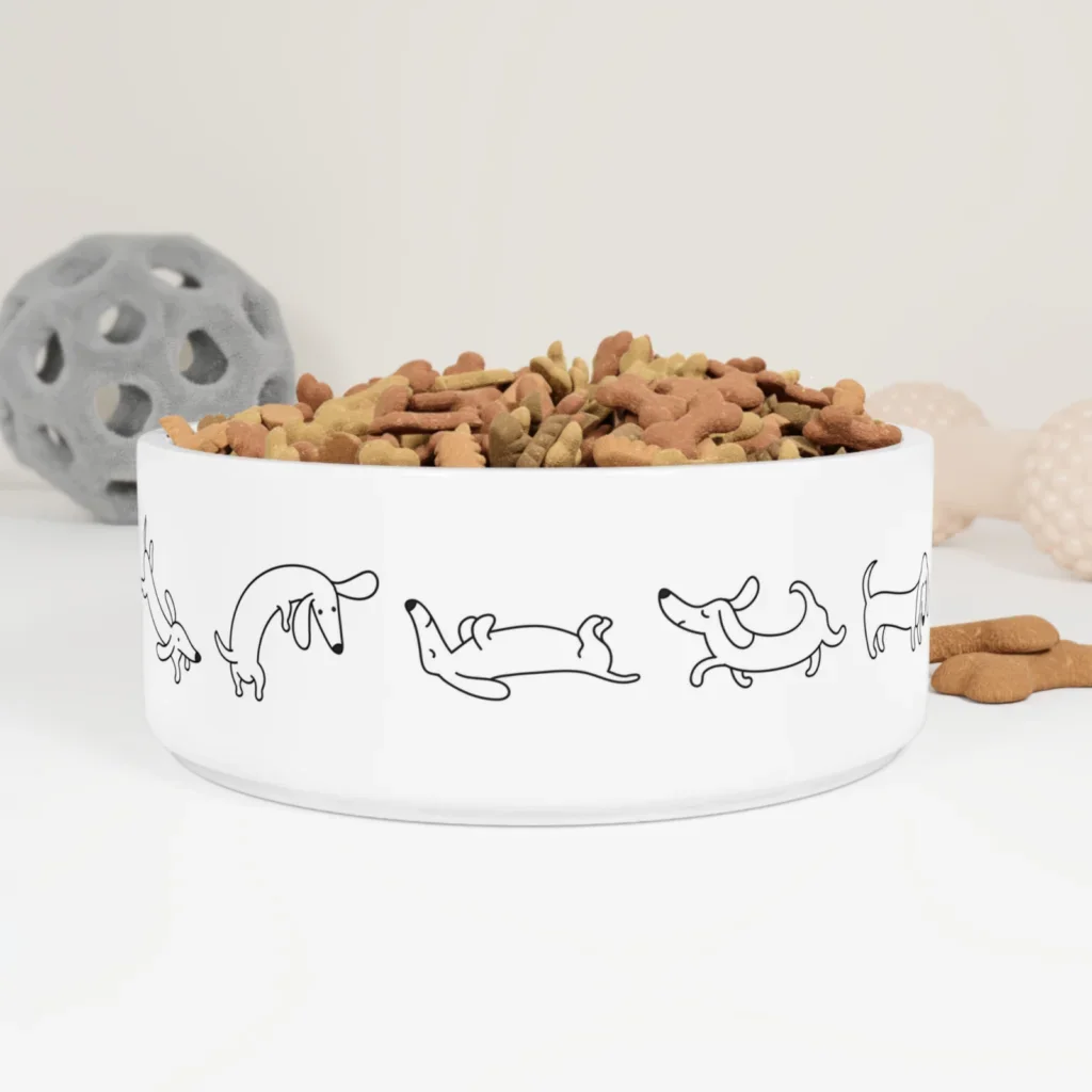 A white dog bowl with cartoon dachshunds is filled with bone-shaped kibble. In the background are a gray toy ball and beige chew toy.