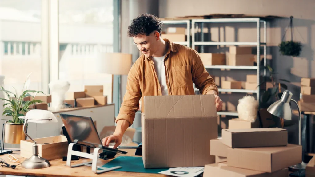A man in a brown shirt happily packs a large cardboard box in a modern office filled with stacked boxes, a computer, and plants.