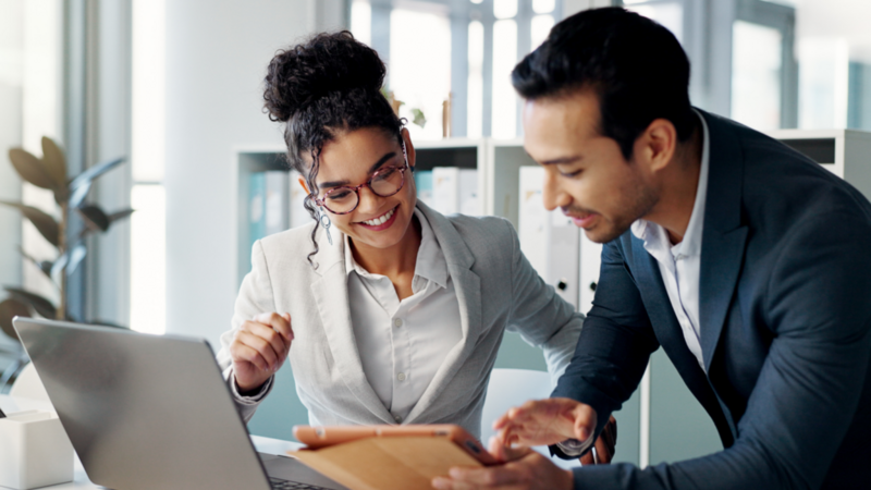 Two colleagues in a bright office, smiling and collaborating over a tablet.