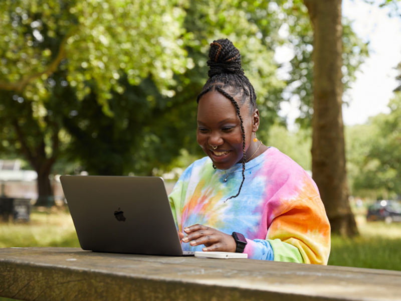A young woman in a tie dye sweatshirt sits at a table in the park, focused on her laptop while enjoying the outdoors.