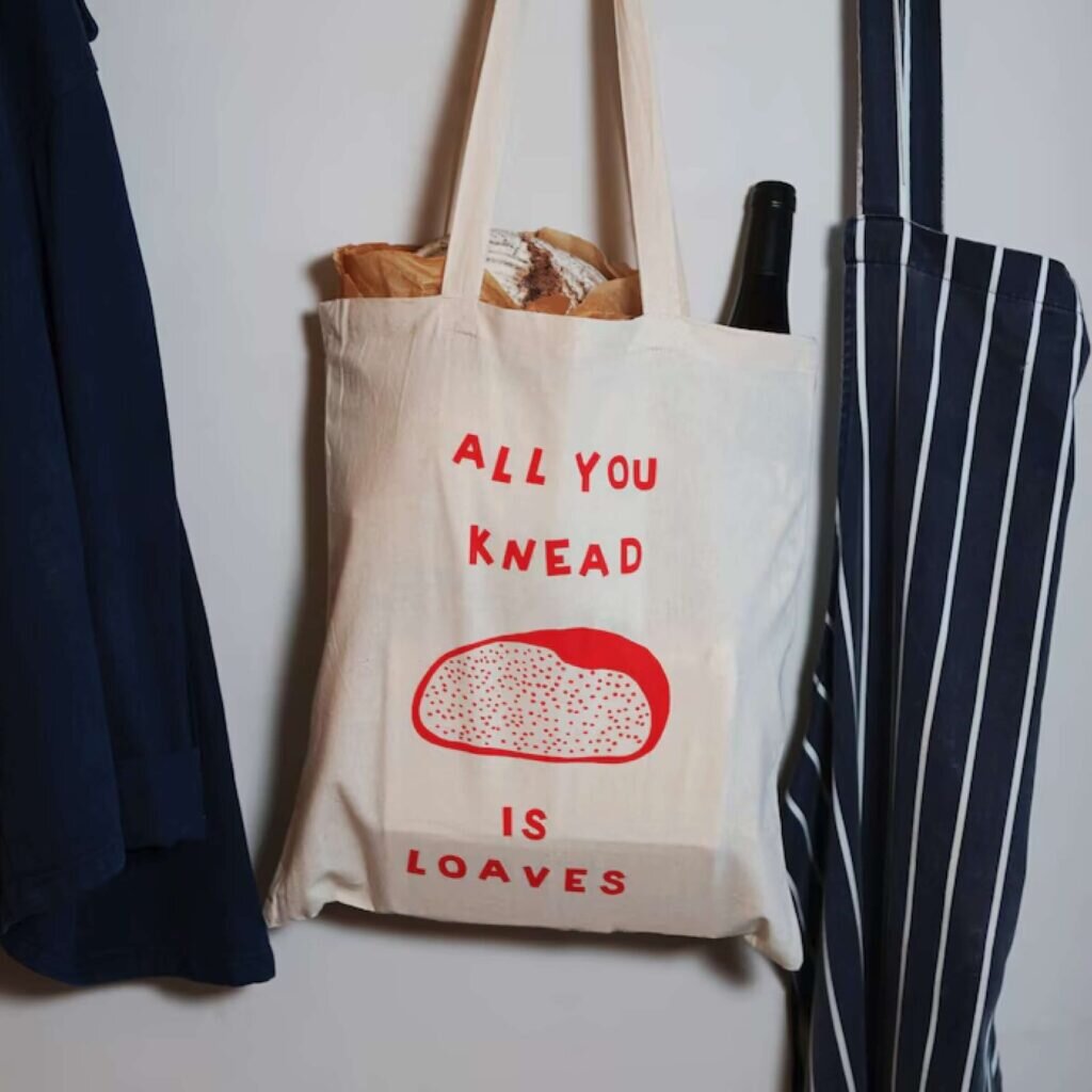 A tote bag with an image of a loaf of bread and the text “All you knead is loaves.”