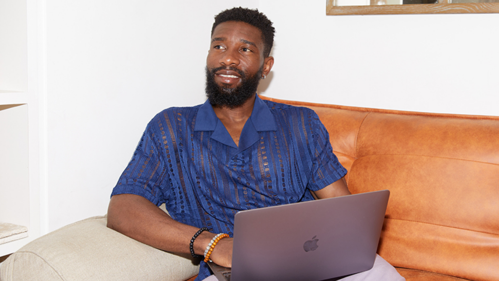 A smiling man in a blue shirt sits on an orange leather couch with a laptop.