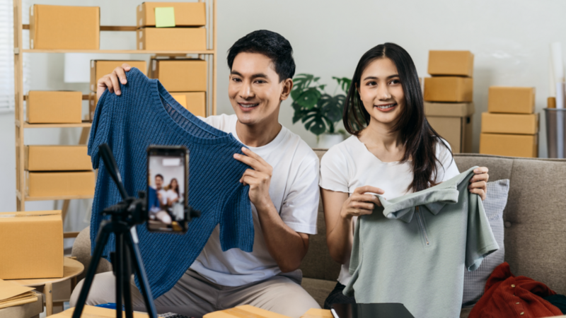 A smiling man and woman hold shirts while live-streaming on a smartphone in a room filled with cardboard boxes.