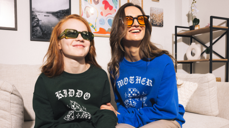 Two women sit on a couch, smiling and wearing sunglasses.