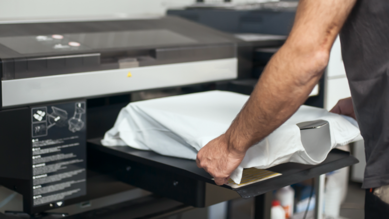 A person places a white t-shirt into a large DTG printing machine, preparing for printing.