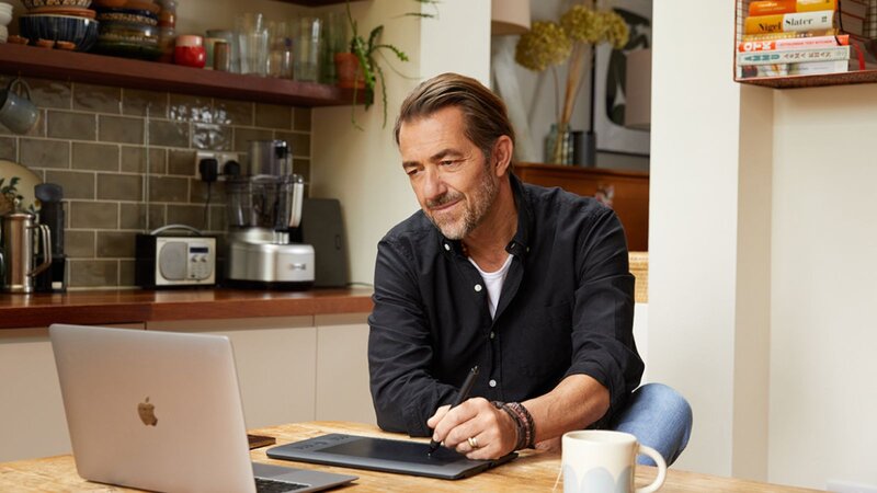 A focused man sitting at a table with a laptop to search for graphic design tips, and a graphics tablet in a kitchen setting.