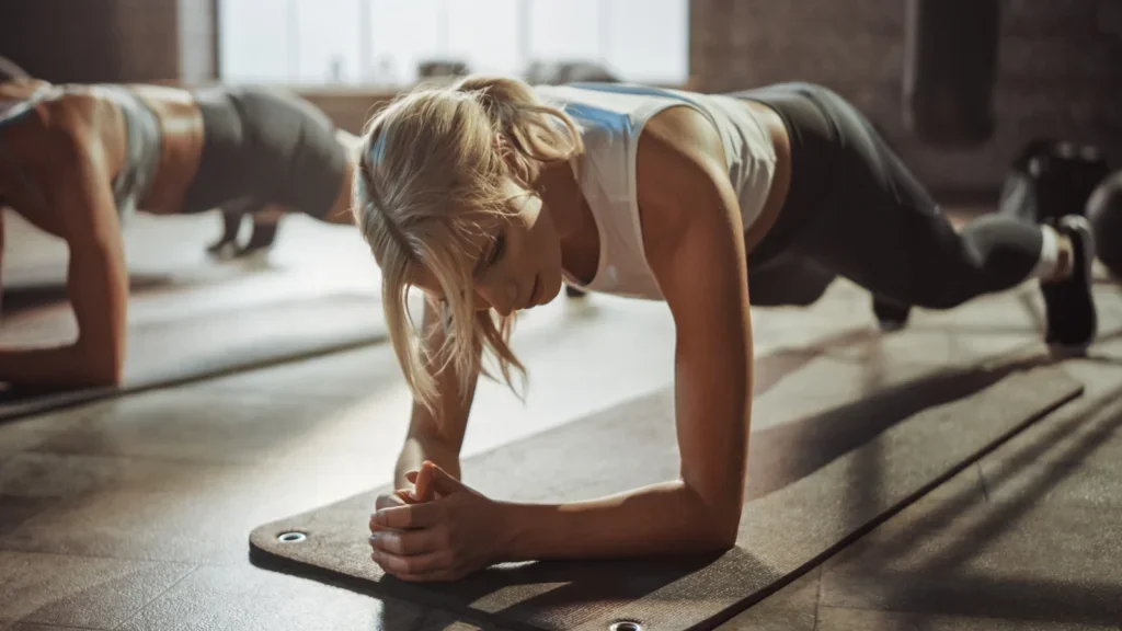 Two women performing push-ups on a fitness mat in a bright, spacious gym environment.