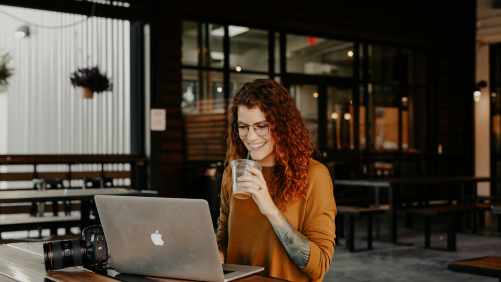 A smiling woman sitting outside in a coffee shop, drinking coffee and using a laptop.