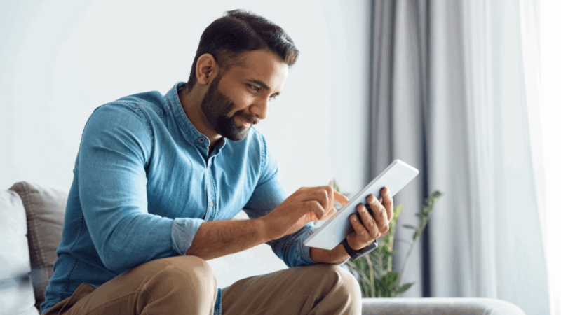 A man in a blue shirt sits on a sofa, smiling and using a tablet to find out if print-on-demand is profitable.
