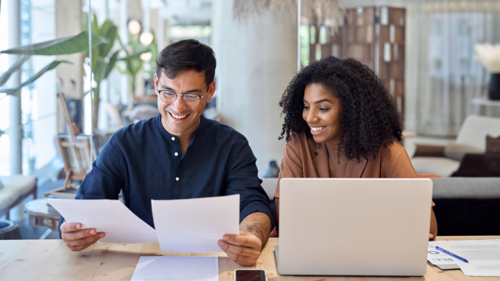 Two individuals seated at a table, reviewing paperwork and using a laptop for their work.